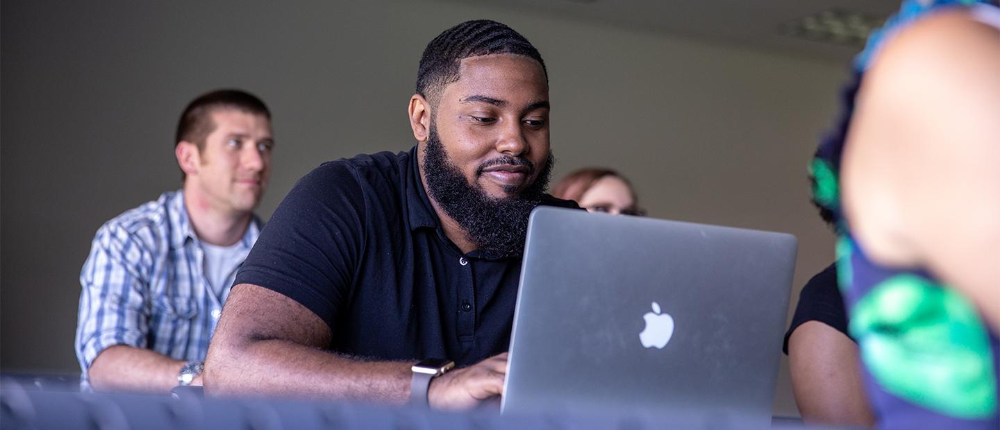 Image of a student wearing a black shirt in a classroom looking at his computer.